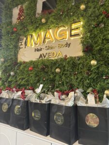 A row of black gift bags with gold logos is displayed in front of a festive green wall with ornaments and a sign reading “THE IMAGE Hair. Skin. Body. AVEDA.”. - The Image Salon & Day Spa, CA