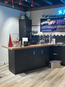 A modern bar setup with black cabinets, a wooden countertop, drink bottles, cups, and holiday decorations, including red ornaments and a small artificial Christmas tree. - The Image Salon & Day Spa, CA