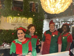 Four people wearing red and green holiday elf costumes stand behind a decorated reception desk with a sign that reads "IMAGE Hair • Skin • Body." Holiday decorations are visible in the background. - The Image Salon & Day Spa, CA