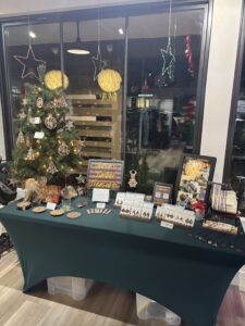 A craft display table with jewelry, ornaments, and wooden decor is set up beside a decorated Christmas tree and a window with hanging star lights. - The Image Salon & Day Spa, CA