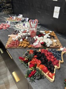 A holiday-themed charcuterie board with cookies, candy, and marshmallows, and a tree-shaped tray filled with assorted fresh berries, set on a dark countertop. - The Image Salon & Day Spa, CA