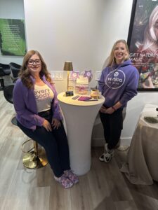 Two women stand and sit by a tall round table displaying wellness products and informational materials in a salon setting, both wearing branded purple clothing. - The Image Salon & Day Spa, CA