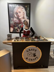 A woman in festive attire stands behind a Crave & Craft food station, with assorted ingredients on display, in front of an Aveda poster. - The Image Salon & Day Spa, CA