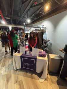 A woman stands behind a vendor table with a purple "Squares Beauty" tablecloth at an indoor market event. Products and informational signs are displayed on the table. - The Image Salon & Day Spa, CA