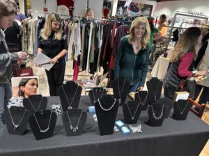 Two women stand behind a table displaying various necklaces and jewelry at an indoor market, with clothing racks and shoppers visible in the background. - The Image Salon & Day Spa, CA