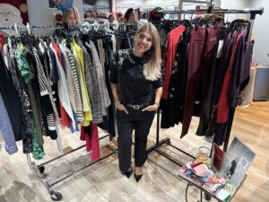 A woman stands smiling in front of two racks of assorted clothing in a retail setting. A small table with flyers, a business card holder, and a drink is nearby. - The Image Salon & Day Spa, CA
